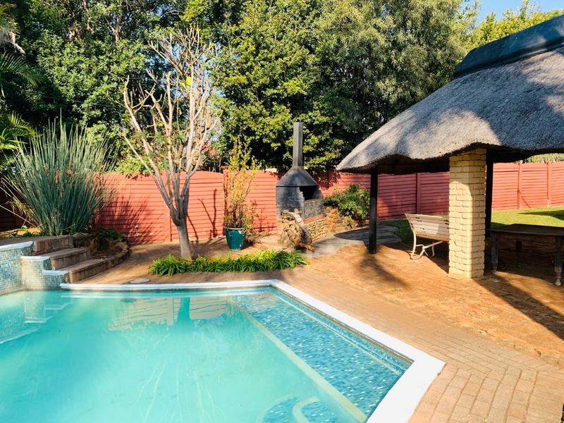 Outdoor entertainment area featuring a swimming pool, a thatched lapa, and a built-in brick braai, set within a private garden with perimeter fencing.