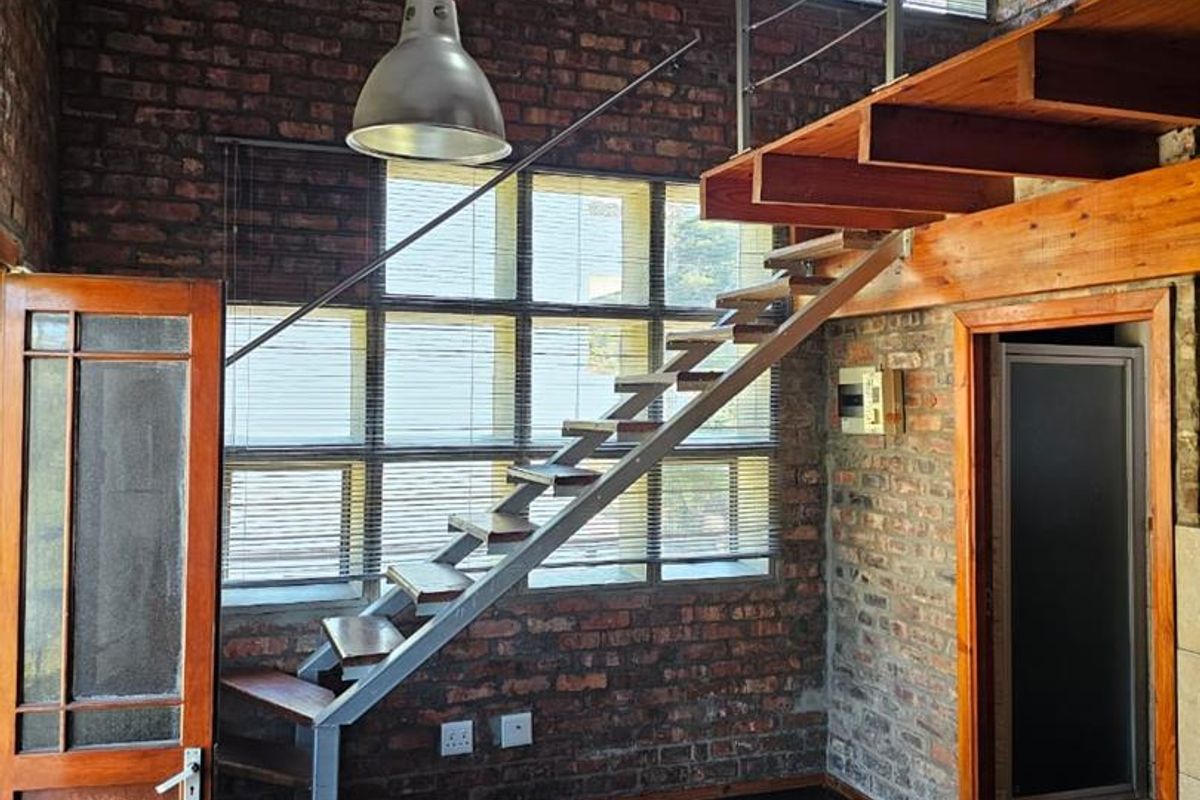 Spacious living area featuring industrial-style exposed brick walls, vaulted ceilings, and a prominent timber staircase leading to a mezzanine level.