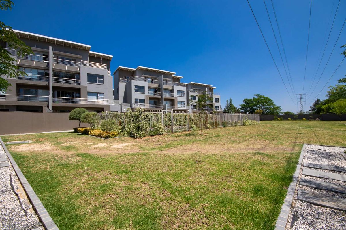 A view of the apartment complex exterior featuring a well-maintained lawn, landscaped garden area, and secure perimeter fencing.