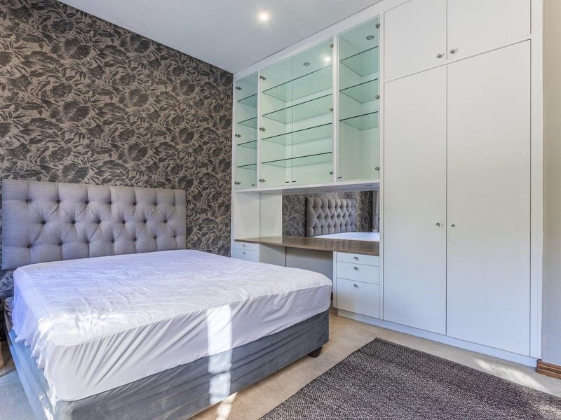 A well-maintained bedroom featuring extensive built-in cabinetry, glass display shelving, a dedicated desk area, and patterned feature wallpaper.