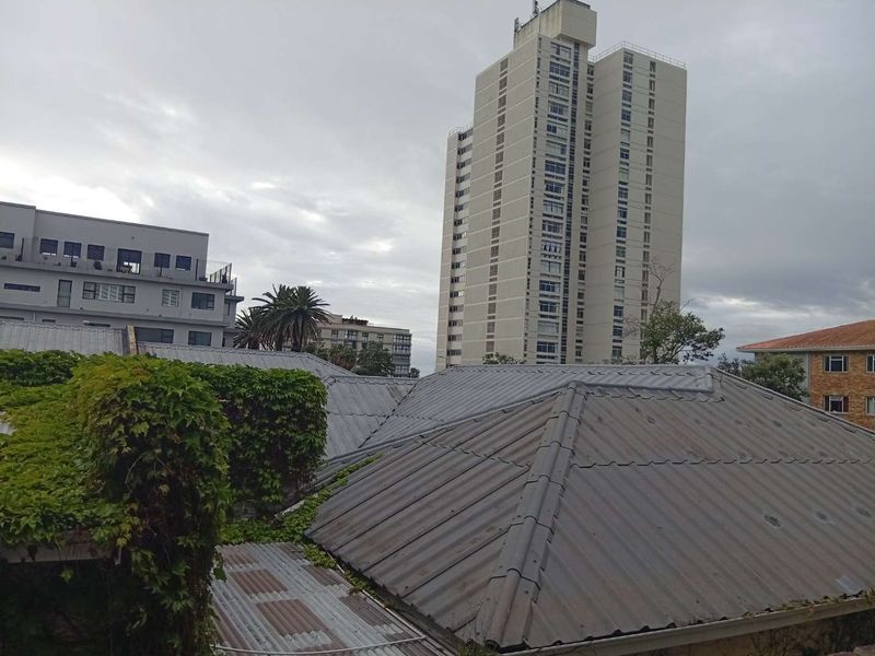 An elevated view overlooking residential rooftops with corrugated metal finishes, featuring a prominent high-rise apartment building in the background under an overcast sky.