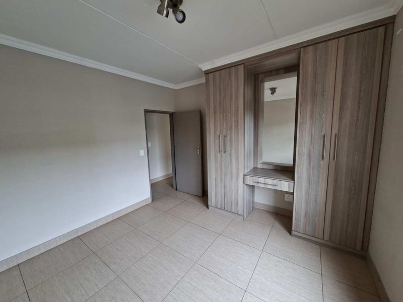 A clean, empty bedroom featuring neutral tiled flooring, coving, and extensive built-in wooden cupboards with an integrated vanity mirror.