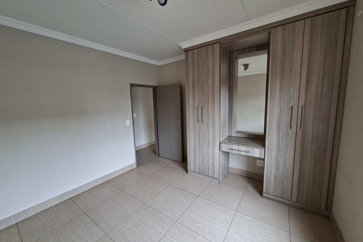 A clean, empty bedroom featuring neutral tiled flooring, coving, and extensive built-in wooden cupboards with an integrated vanity mirror.