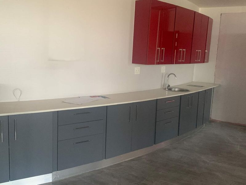 A modern kitchenette featuring grey base cabinets, red wall-mounted cupboards, a stone countertop, and a stainless steel sink in an unfinished office space.