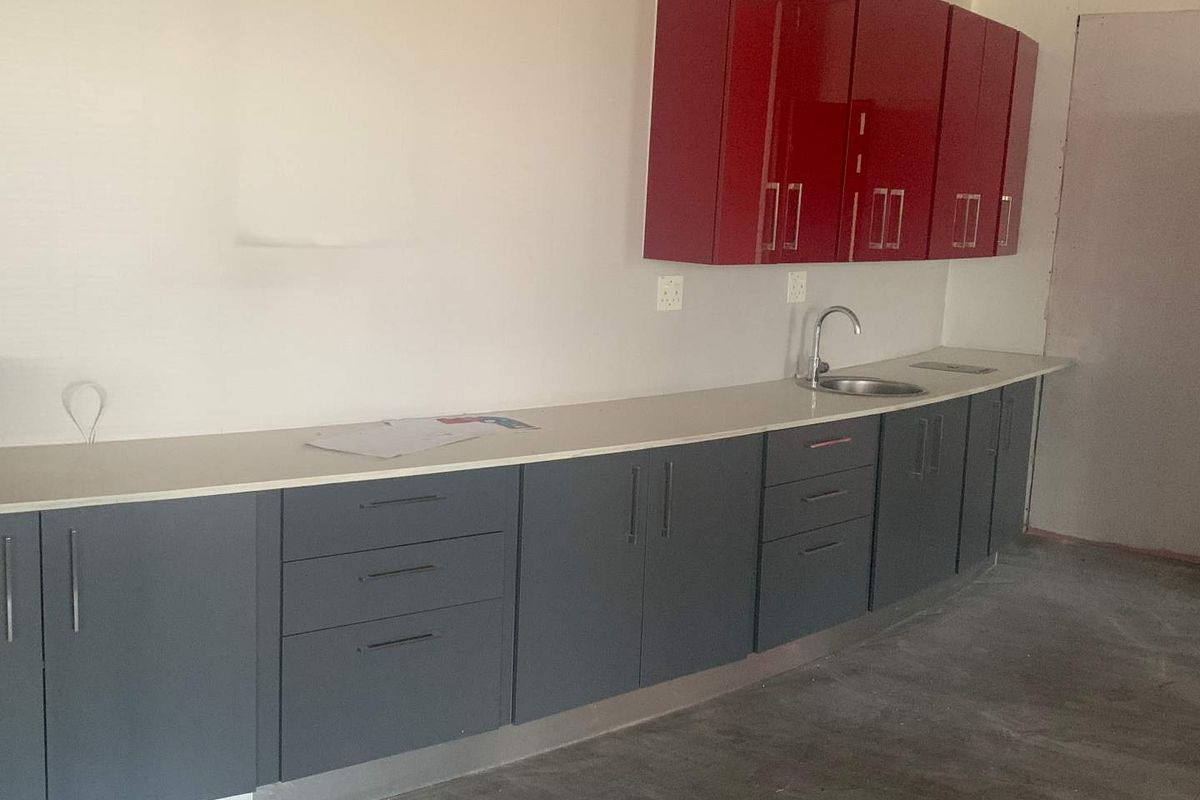A modern kitchenette featuring grey base cabinets, red wall-mounted cupboards, a stone countertop, and a stainless steel sink in an unfinished office space.