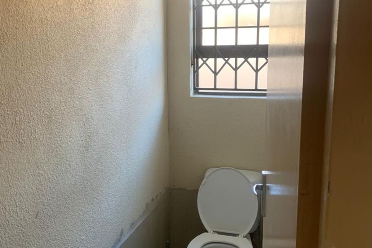 A compact bathroom featuring a standard toilet, tiled flooring, and a window with security bars, currently requiring cosmetic updates to the wall finishes.
