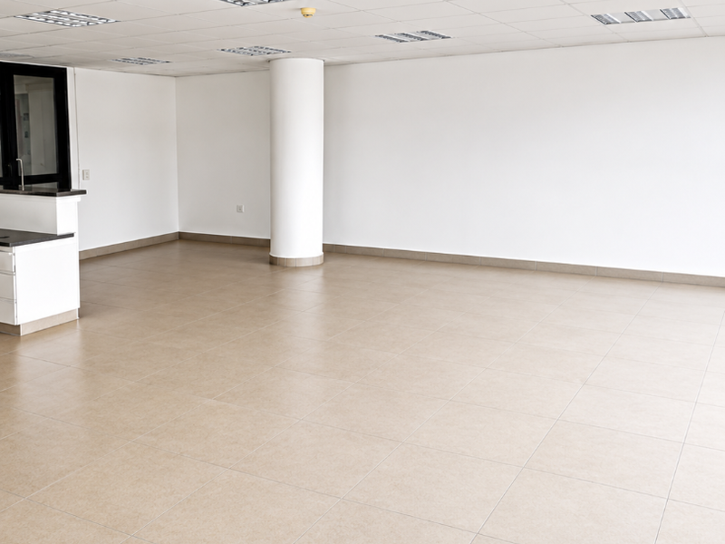 A spacious, open-plan office area featuring neutral ceramic floor tiles, a suspended ceiling with recessed lighting, and a central structural column.
