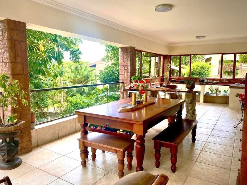 Spacious covered patio featuring tiled flooring, stone-clad pillars, and a glass balustrade, offering a seamless transition to the lush garden view.