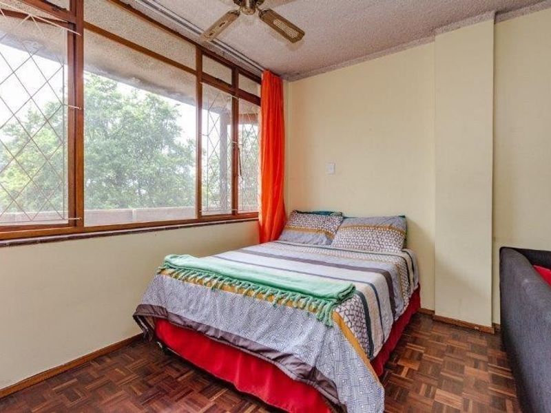 A bedroom featuring original parquet flooring, a ceiling fan, and large windows with security bars, offering potential for modernization.