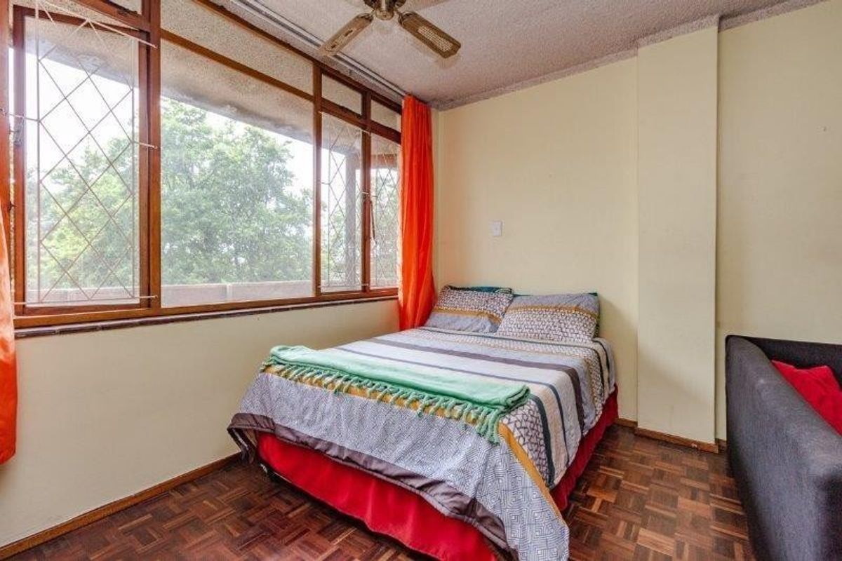 A bedroom featuring original parquet flooring, a ceiling fan, and large windows with security bars, offering potential for modernization.