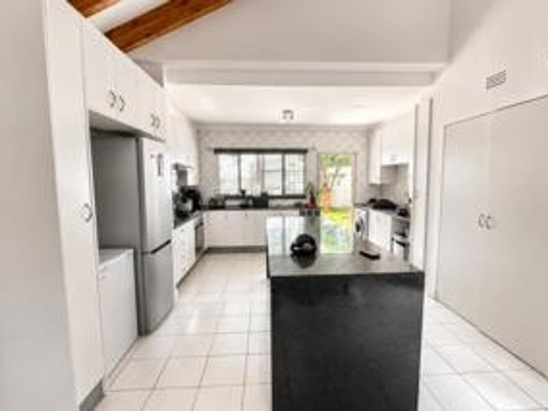 A bright, modern kitchen featuring a central island with a dark granite countertop, ample built-in cabinetry, tiled flooring, and exposed timber ceiling beams.