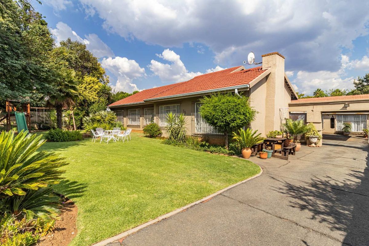 A well-maintained rear exterior view of the house featuring a spacious lawn, paved driveway, and a tiled roof with a chimney.