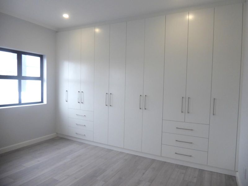 A bright, modern bedroom featuring extensive floor-to-ceiling built-in cupboards, light-toned laminate flooring, and a clean, minimalist aesthetic.