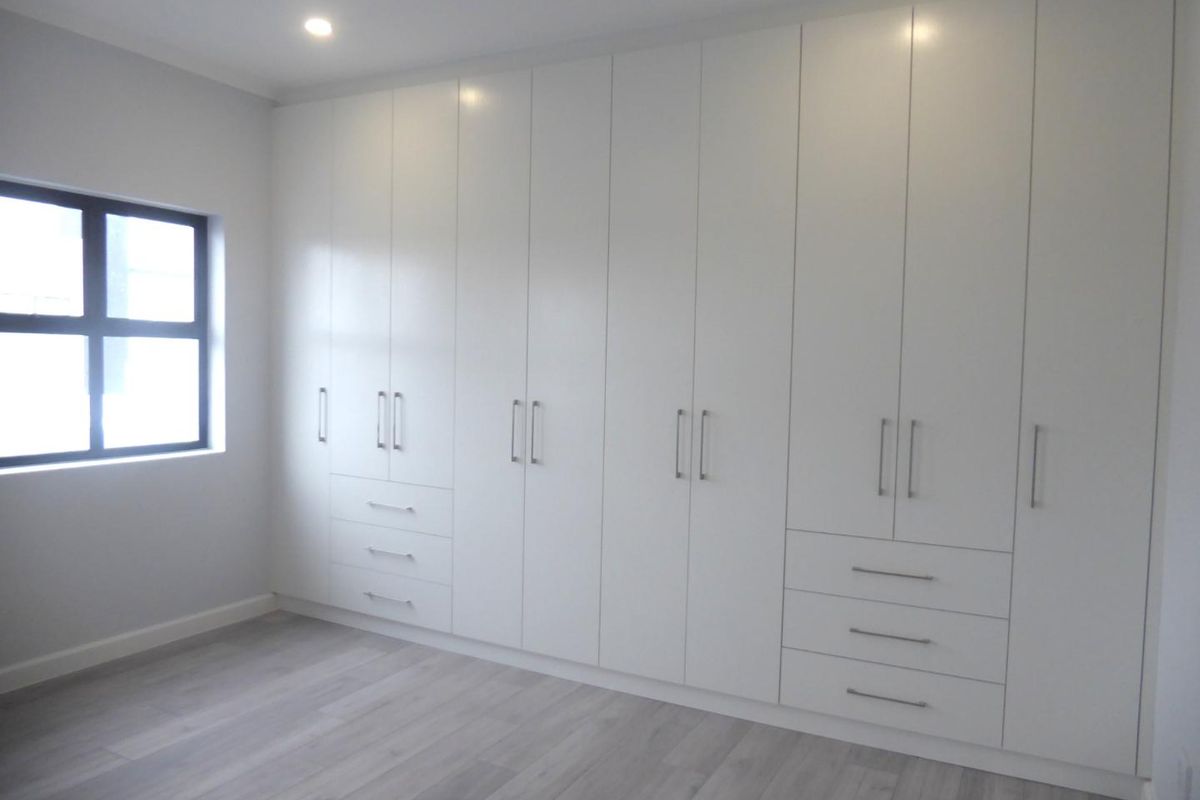 A bright, modern bedroom featuring extensive floor-to-ceiling built-in cupboards, light-toned laminate flooring, and a clean, minimalist aesthetic.