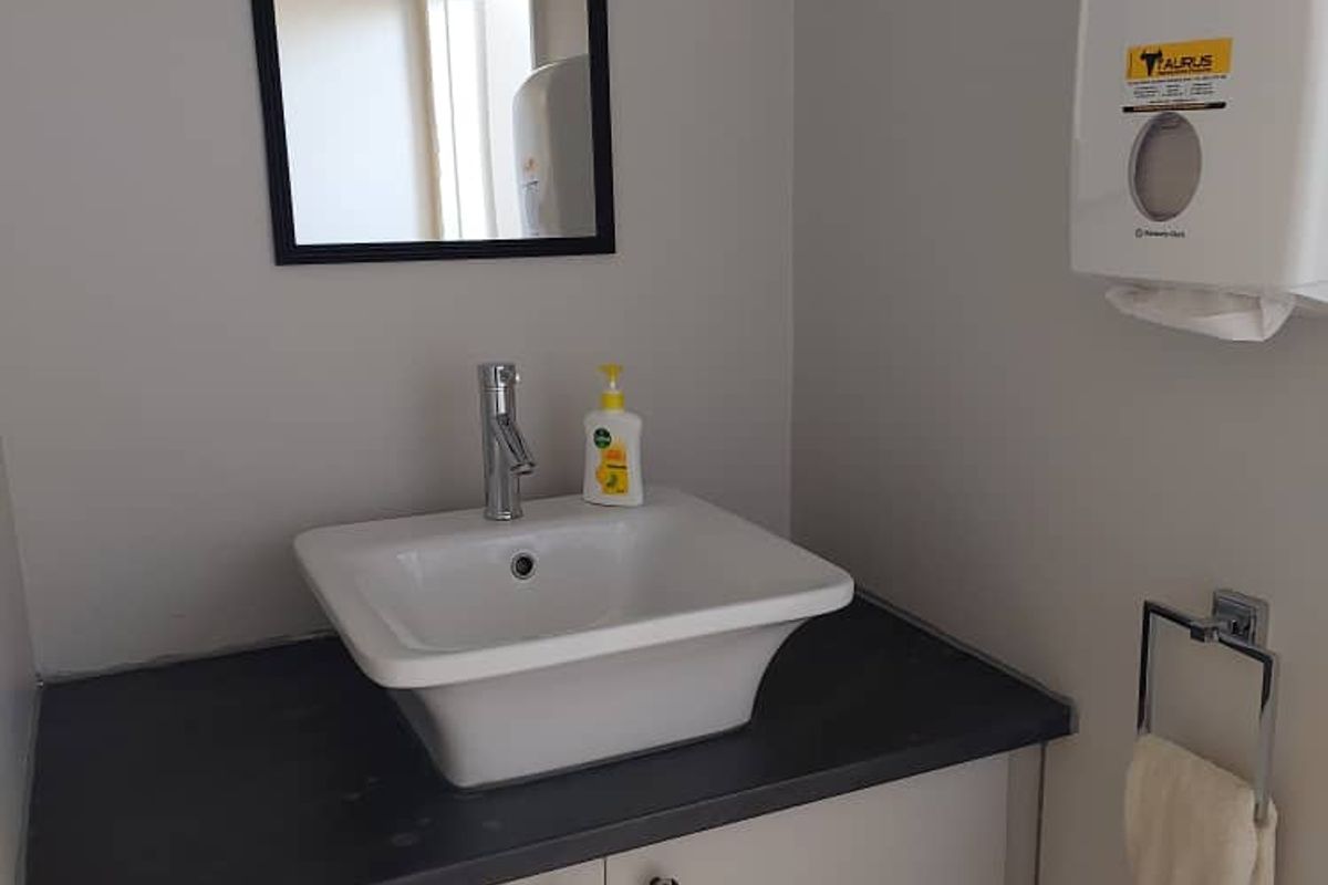 A functional office restroom vanity area featuring a modern vessel sink, dark countertop, storage cabinetry, wall-mounted mirror, and paper towel dispenser.