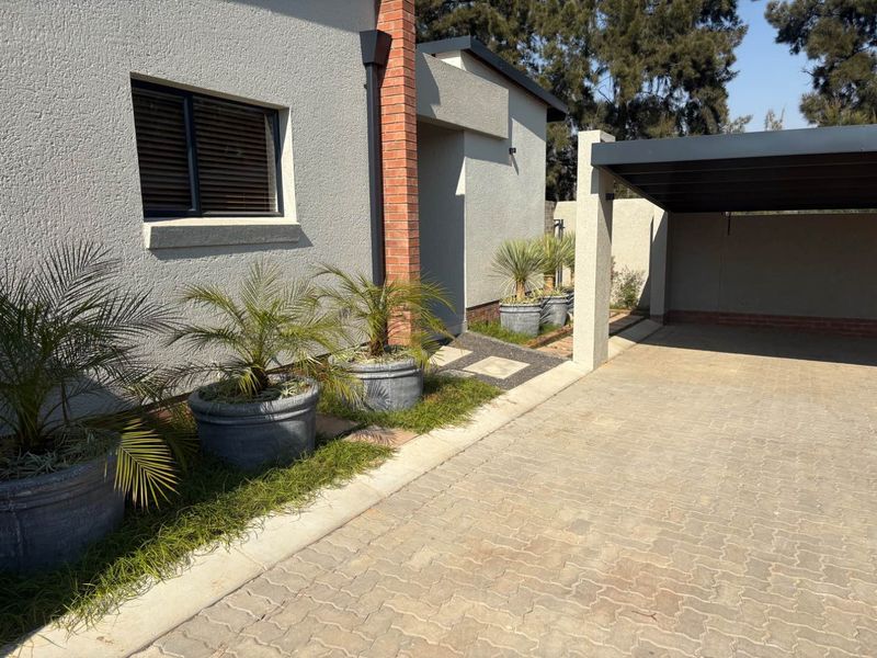 Front exterior view of the apartment featuring a paved driveway, covered carport, and a well-maintained facade with brick detailing.
