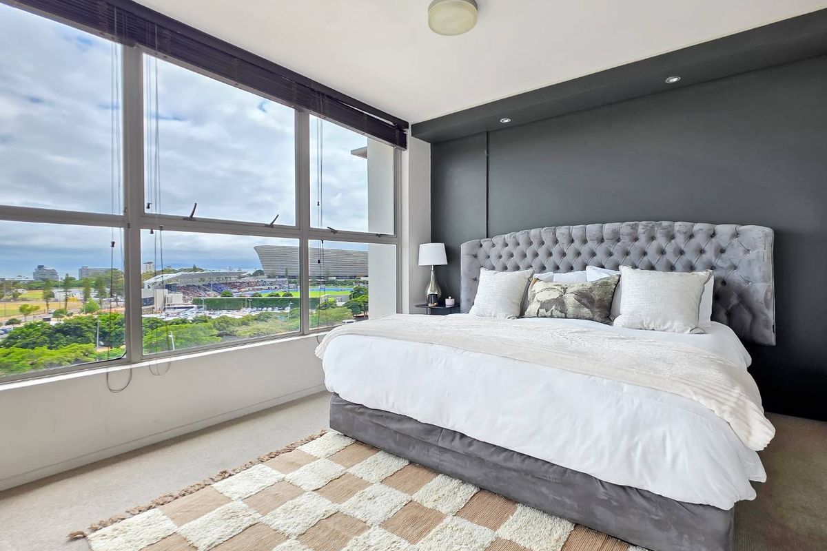A bright, modern bedroom featuring large floor-to-ceiling windows with expansive city and stadium views, complemented by a dark feature wall and neutral carpeting.