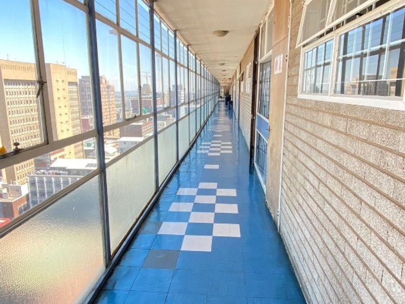 A long, open-air apartment hallway featuring large windows with city views, brick wall finishes, and blue and white tiled flooring.