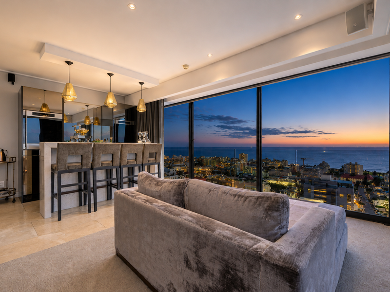 Modern open-plan living area featuring a sleek built-in bar, stone flooring, and expansive floor-to-ceiling windows showcasing panoramic sunset ocean and city views.