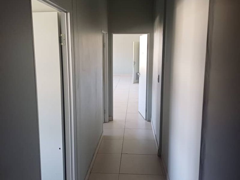 A clean, empty office hallway featuring neutral grey walls, tiled flooring, and a suspended ceiling with recessed lighting.