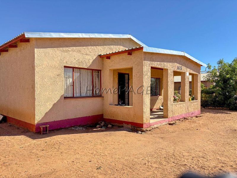 Front elevation of a single-story house featuring a covered porch, metal roofing, and security bars on windows, set on an unpaved lot.