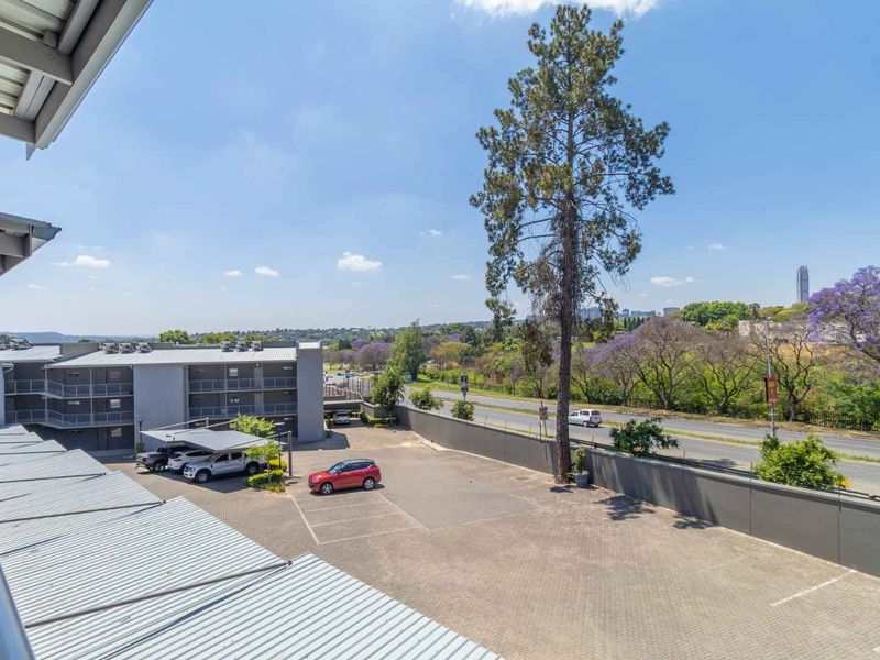 Elevated view overlooking the apartment complex parking area, perimeter wall, and surrounding landscape with mature trees.