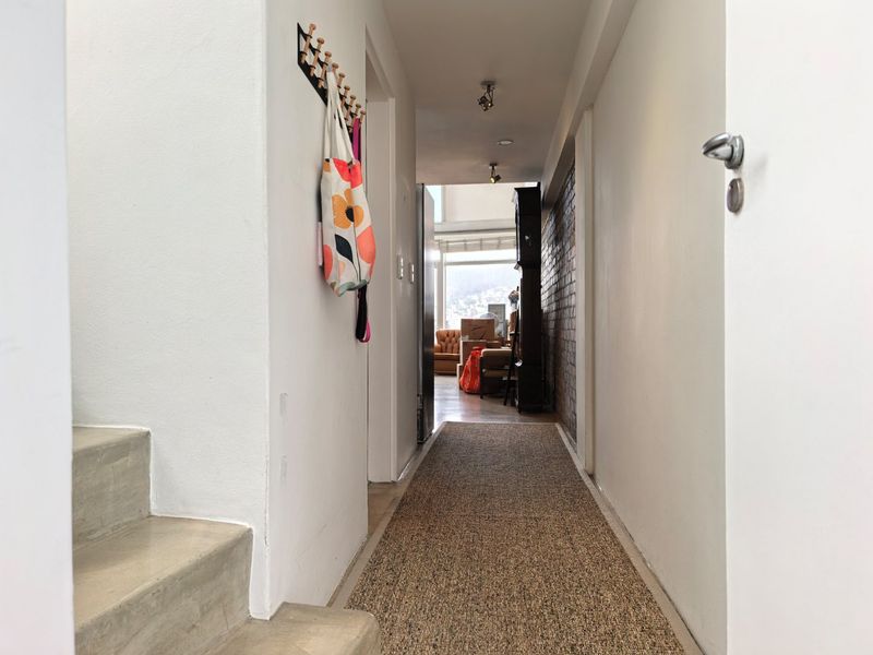 A bright hallway featuring an exposed brick wall and concrete staircase, leading towards an open-plan living area with natural light.