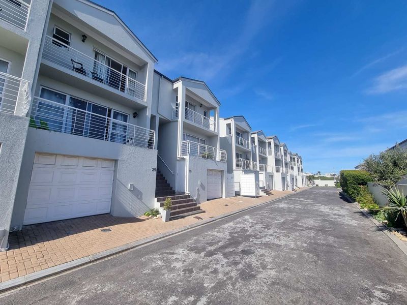 A row of modern townhouse units featuring paved driveways, integrated garages, and upper-level balconies with metal railings under a clear blue sky.