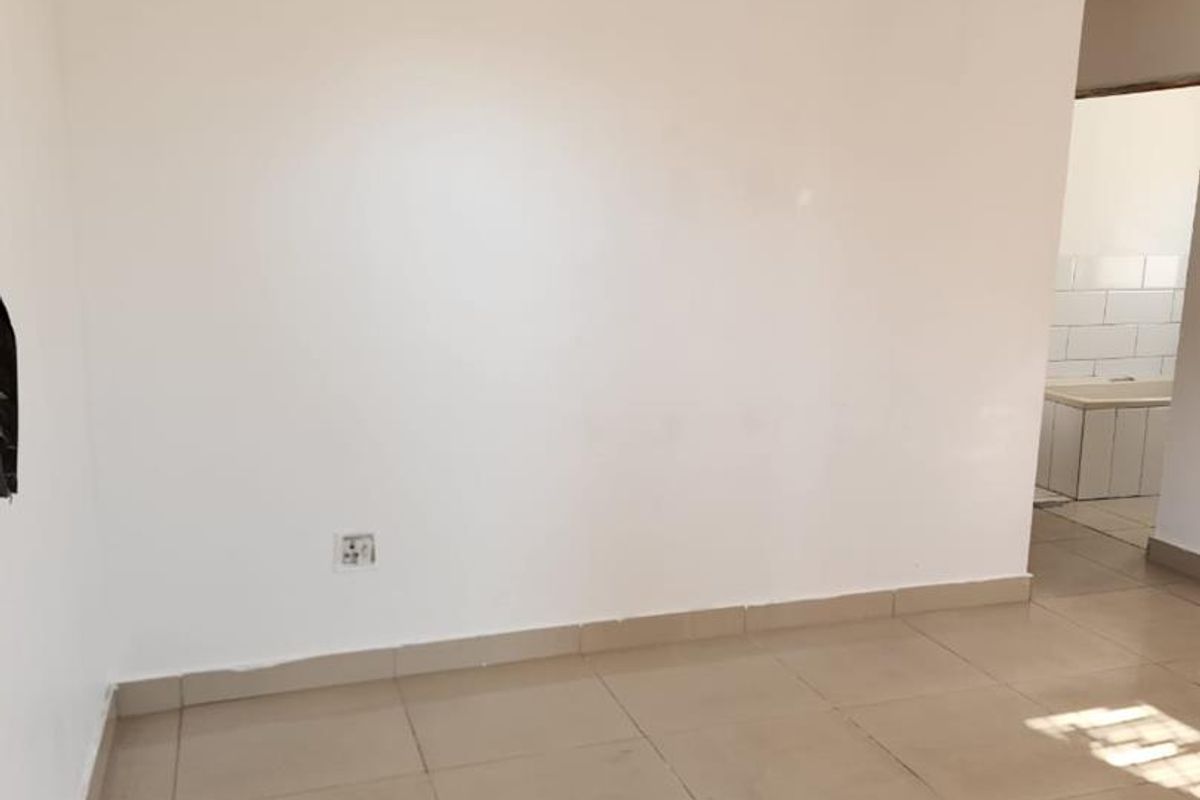 A vacant living area featuring neutral ceramic floor tiles and plain white walls, with a view into an adjacent tiled space.