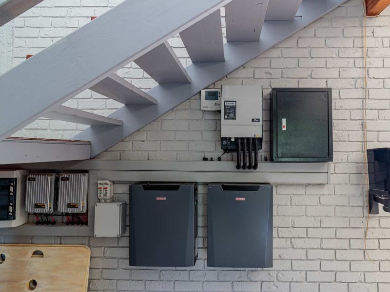 Utility area featuring a solar power inverter and battery storage system mounted on a painted brick wall beneath a staircase.