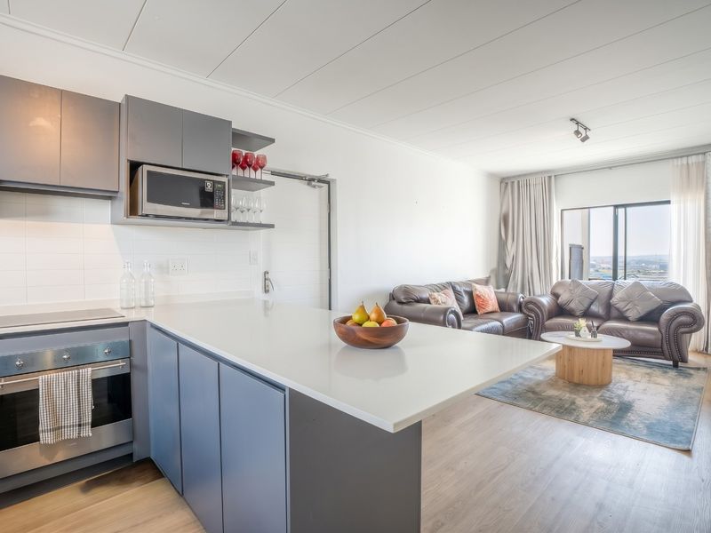 Modern open-plan apartment featuring a sleek kitchen with stone countertops and built-in appliances, flowing seamlessly into a bright living area with large windows.
