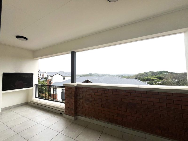 A spacious covered balcony featuring tiled flooring, a built-in braai, and a brick balustrade with metal railing, offering pleasant neighbourhood views.