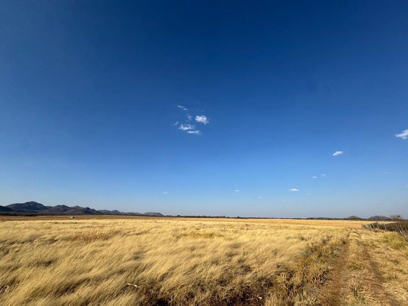 Expansive open farm landscape featuring dry grassland, distant mountain ranges, and a clear blue sky.