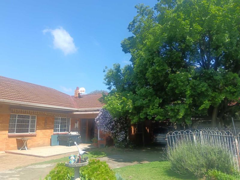 A rear view of a brick house featuring a tiled roof, a paved patio area, and a carport, set within a mature garden.