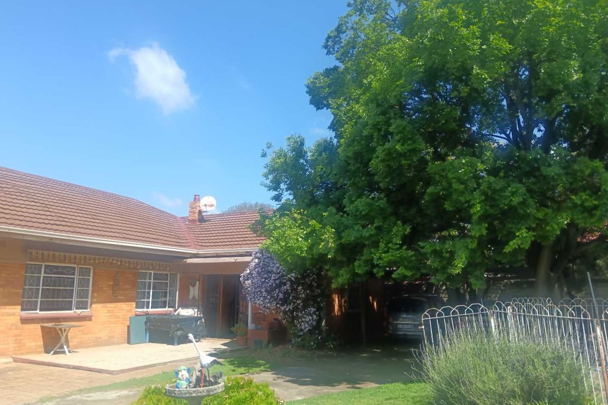 A rear view of a brick house featuring a tiled roof, a paved patio area, and a carport, set within a mature garden.