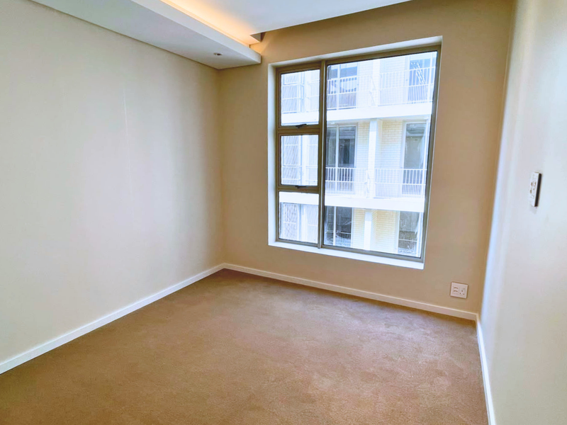 A clean, empty bedroom featuring neutral walls, carpeted flooring, and a large window providing natural light and a view of the adjacent building.