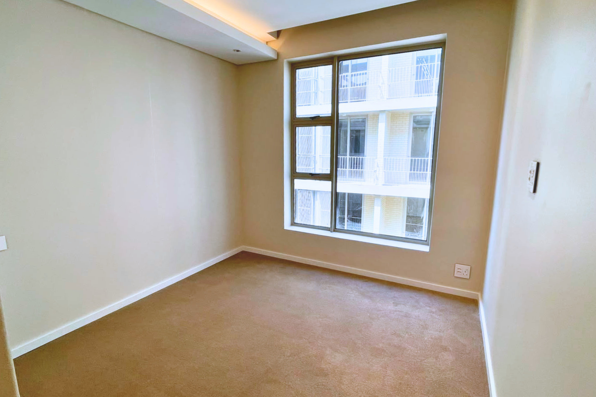 A clean, empty bedroom featuring neutral walls, carpeted flooring, and a large window providing natural light and a view of the adjacent building.