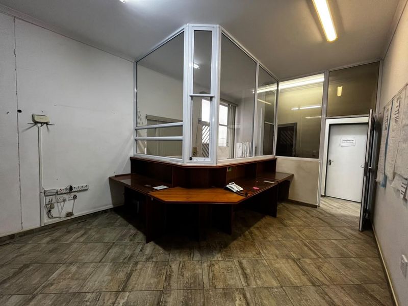 An industrial office space featuring a built-in reception desk, glass partition, tiled flooring, and functional fluorescent lighting.