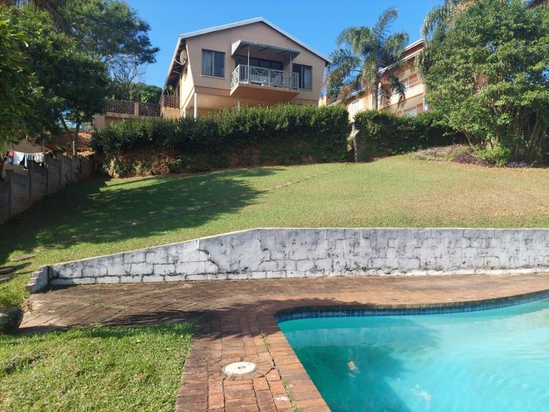 Rear exterior view featuring a swimming pool with brick surround, a tiered lawn, and a multi-level house with a balcony.