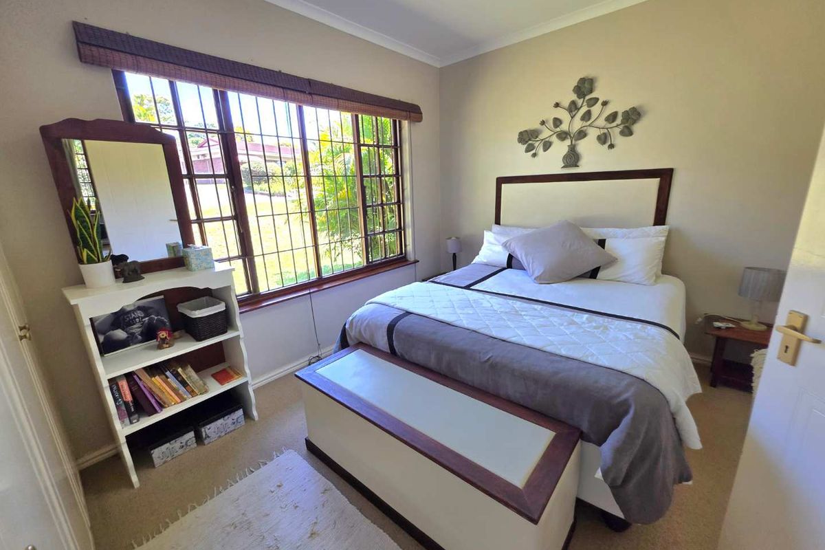 A well-lit bedroom featuring a large window with security bars, neutral wall tones, and carpeted flooring.