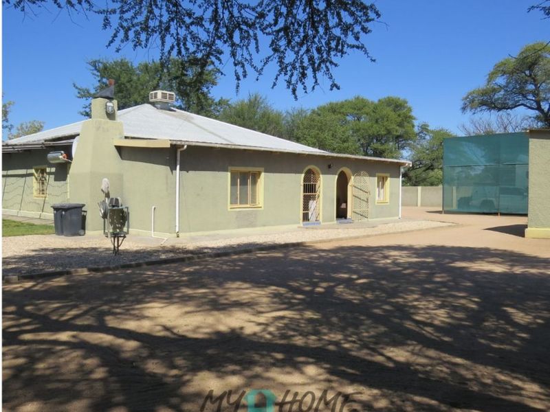 Exterior view of a single-story house featuring a metal roof, arched entrance, and security gates, set on a large plot with gravel driveway.