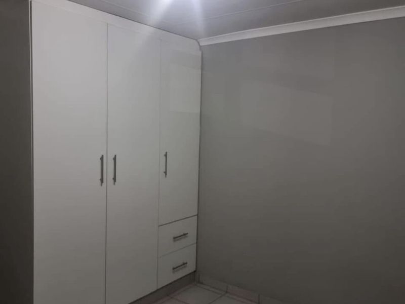 A clean, empty bedroom featuring neutral grey walls, tiled flooring, and modern white built-in cupboards with integrated drawers.