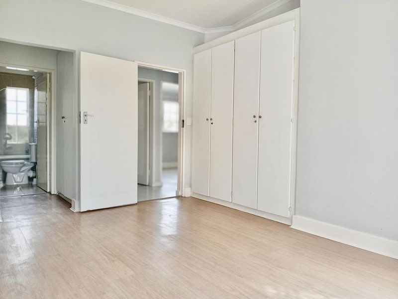 A bright, empty bedroom featuring light-toned laminate flooring, built-in cupboards, and neutral wall finishes, with an adjacent view into a bathroom.