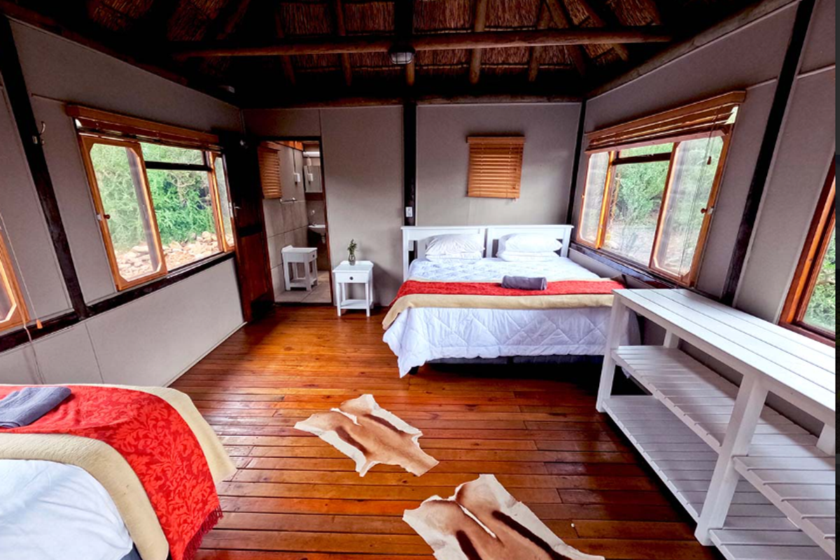 A rustic bedroom featuring a traditional thatched roof, exposed timber beams, wooden flooring, and large windows with natural light, including an en-suite bathroom.