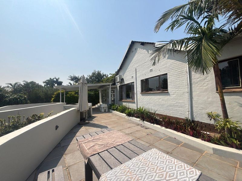 A spacious rear patio featuring stone paving, landscaped garden beds, and a white brick exterior wall with an integrated air conditioning unit.