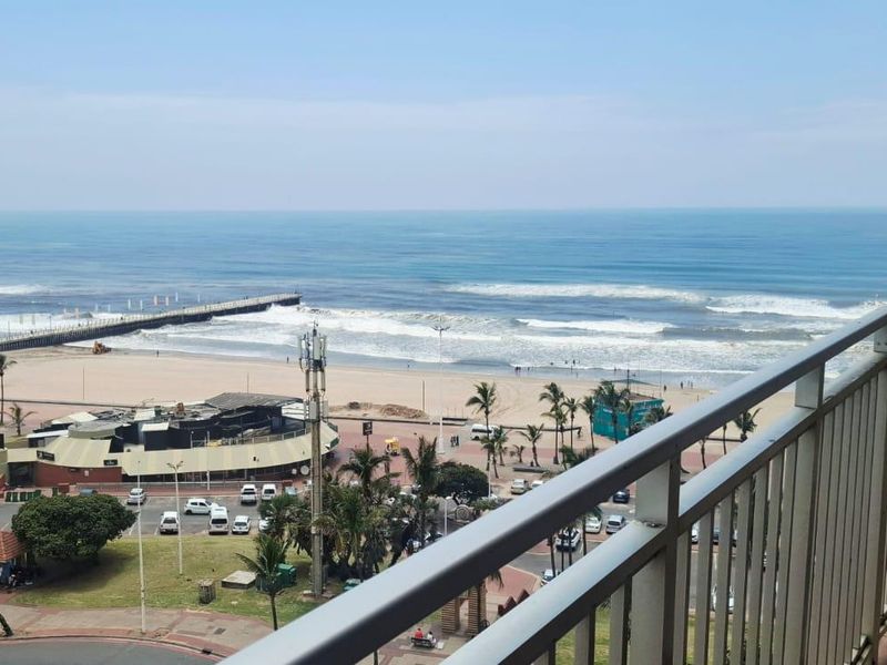 A balcony featuring a metal balustrade and tiled flooring, offering an expansive, elevated view of the beach, ocean, and coastal promenade.
