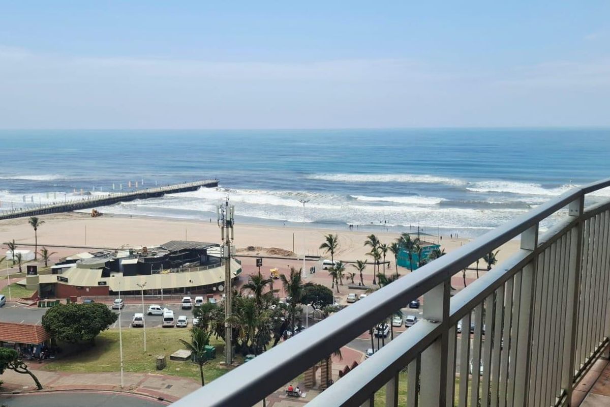 A balcony featuring a metal balustrade and tiled flooring, offering an expansive, elevated view of the beach, ocean, and coastal promenade.