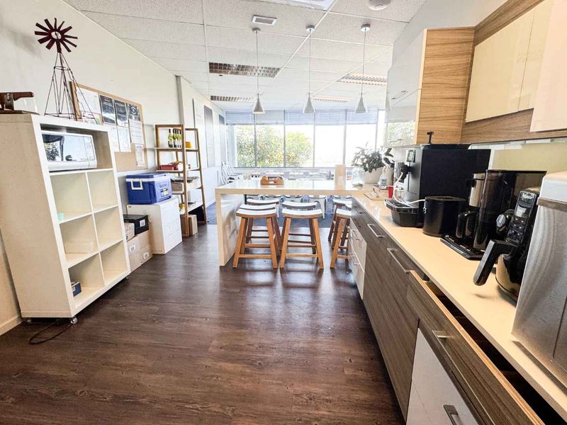 A functional office kitchenette featuring modern laminate cabinetry, a long breakfast bar, and large windows providing ample natural light.