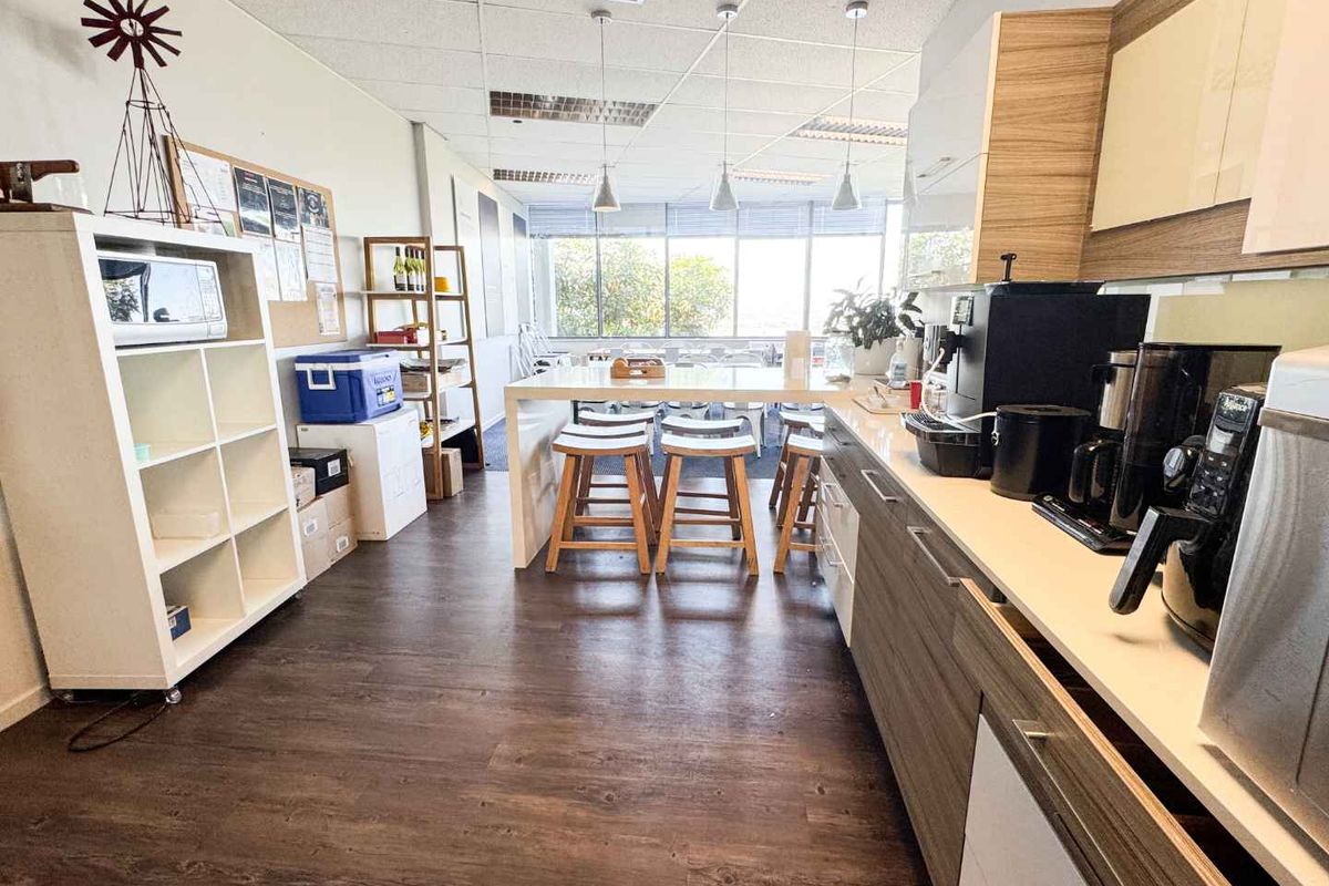 A functional office kitchenette featuring modern laminate cabinetry, a long breakfast bar, and large windows providing ample natural light.