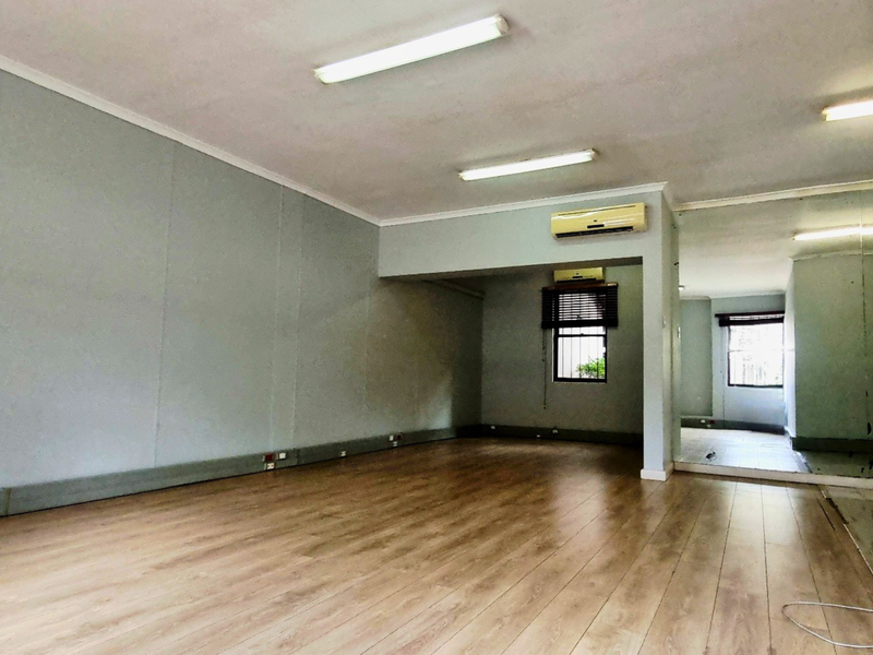 An empty office space featuring laminate flooring, fluorescent lighting, a wall-mounted air conditioning unit, and a large mirrored wall section.